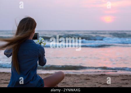 Eine Frau sitzt am Meer und fotografiert mit ihrem Smartphone vor dem Hintergrund der untergehenden Sonne eine Plumeria-Blume. Die Szene ist ruhig und pe Stockfoto