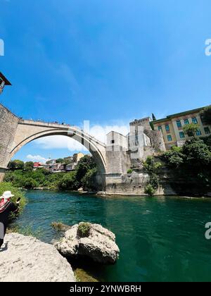 Eine historische Steinbrücke führt anmutig über einen pulsierenden türkisfarbenen Fluss, umgeben von üppigem Grün und alter Architektur unter einem klaren blauen Himmel. Stockfoto