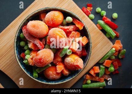 Eine Nahaufnahme von buntem gefrorenem Gemüse, einschließlich Erbsen, Hülsenfrüchten, Tomaten, Paprika, und Karotten, in einer Schüssel auf dunkelblauem Hintergrund. Stockfoto