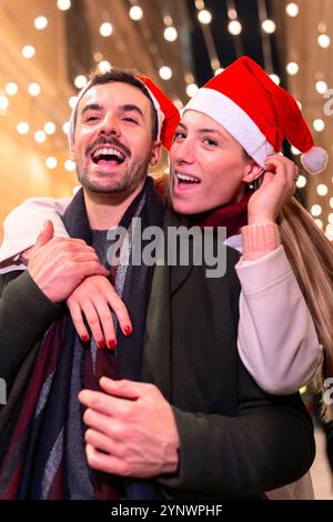 Lachendes Paar mit Weihnachtsmützen auf festlicher Stadtstraße mit Weihnachtsbeleuchtung Stockfoto