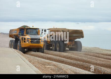 Zwei Volvo A40E Kipper fahren einander vorbei und transportieren Schindeln am Sandgate Beach in der Nähe von Folkestone, Großbritannien. Ein Lkw hat seine Ladung zum Strand geliefert und kehrt zum Nachfüllen zurück. Der Schindel wird jedes Jahr recycelt und bewegt den Schindel entlang der Küste von Ost nach West, um Überschwemmungen und Küstenerosionen zwischen Folkestone und Hythe zu reduzieren. Stockfoto