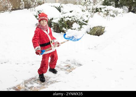 Der Junge trägt einen Weihnachtsmann-Anzug und räumt nach einem starken Schneesturm die Straße zum Haus und zum Hof fleißig vom Schnee ab, hält eine Schaufel in h Stockfoto