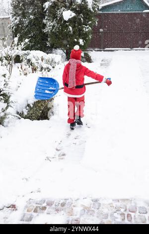 Unerkennbares Kind in einem roten Santi-Kostüm mit großer Schaufel räumt nach einem starken Schneesturm die Straße vom Schnee ab. Wintersaison, schlechtes Wetter, Kälte, Stockfoto