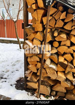 Ein Stapel sauber angeordneten Feuerholzes vor einem roten Zaun in einem schneebedeckten Hinterhof, neben einem blattlosen Baum, der das Wesen der Wintervorbereitung einfängt Stockfoto