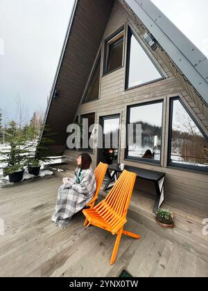Eine ruhige Winterszene mit einer modernen Kabine mit A-Rahmen und einer Person, die einen gemütlichen Sitz auf dem Deck genießt. Die Lage ist friedlich, mit Schnee und A Stockfoto