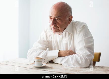 Ein älterer Mann sitzt nachdenklich an einem Holztisch, in einem weißen Hemd gekleidet. Er blickt in die Ferne, eine kleine Tasse Kaffee neben ihm Stockfoto