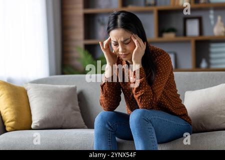Asiatische Frau, die Kopfschmerzen hat und zu Hause auf der Couch sitzt. Die Schläfen mit den Fingern drücken, die gestresst und unbequem aussehen. Freizeitkleidung, Inneneinrichtung, Spannungsgefühl und Ermüdung. Stockfoto