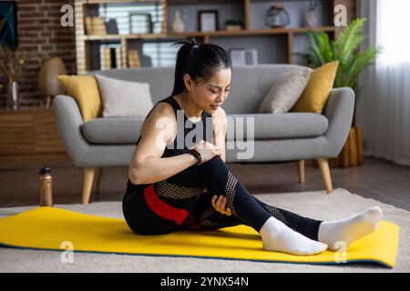 Asiatische Frau verletzt, während sie Fitness und Yoga zu Hause macht, Frau strapazierte Beinmuskulatur, Massage, Muskelschmerzen, sitzend auf Sportmatte im Wohnzimmer in der Wohnung. Stockfoto