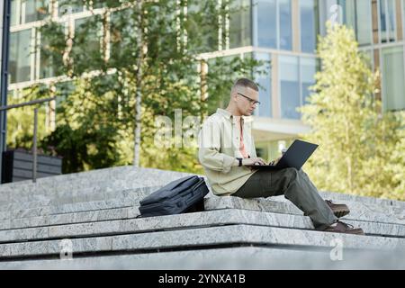 Eine vollständige Aufnahme eines konzentrierten männlichen IT-Programmierers in lässiger Kleidung mit Laptop, um Code zu studieren oder zu schreiben, der auf Betonstufen sitzt und als nächstes den Rucksack liegt Stockfoto