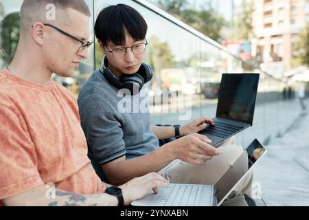 Mittelgroße Aufnahme eines asiatischen Geschäftsmannes mit Kopfhörern, der auf den Laptop-Bildschirm zeigt und mit einem Kollegen am Projekt arbeitet, während er im Cit auf dem Bürgersteig sitzt Stockfoto