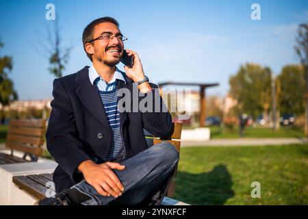 Geschäftsmann im Mantel mit Brille, der an einem sonnigen Herbsttag auf einer Bank im Park sitzt. Stockfoto