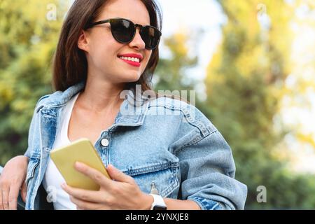 Stilvolle weiße Frau mit Jeansjacke und Sonnenbrille, die im Freien mit einem Smartphone posiert, in einer urbanen Umgebung mit verschwommenem Grün im Hintergrund Stockfoto