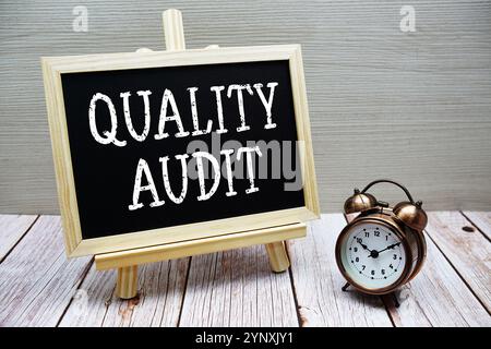 Text der Qualitätsprüfung auf Tafel geschrieben mit Wecker auf hölzernem Hintergrund Stockfoto