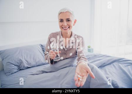 Lächelnde ältere Frau in Nachtwäsche, die ein Glas Wasser anbietet, während sie im Morgenlicht bequem im Bett sitzt Stockfoto