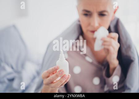 Ältere Frau im Schlafanzug, die Nasenspray während des Schlafens im Bett während einer ruhigen Morgenroutine untersucht Stockfoto