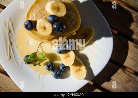 Ein Teller flauschiger Pfannkuchen mit Sirup, belegt mit frischen Bananenscheiben und Heidelbeeren. Diese Frühstücksszene wurde im warmen Morgenlicht aufgenommen Stockfoto