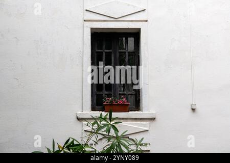 Ein Vintage-schwarzes Fenster, das von einer weißen strukturierten Wand eingerahmt wird und mit einem Übertopf aus leuchtenden rosa Blumen geschmückt ist. Stockfoto