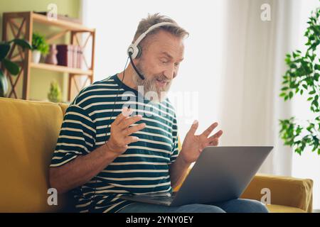 Glücklicher älterer Mann, der zu Hause mit aufgesetztem Headset ein entspanntes Gespräch genießt und bequem in einem gemütlichen Wohnzimmer sitzt. Stockfoto