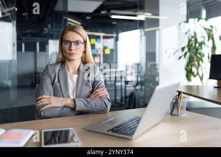 Reife Geschäftsfrau mit Brille in einem eleganten Büroumfeld. Sie strahlt Vertrauen und Autorität aus. Laptop und Telefon auf dem Schreibtisch sorgen für Produktivität und moderne Arbeitsdynamik. Stockfoto