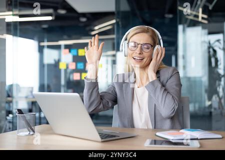 Reife Frau Geschäftsfrau, die sich glücklich fühlt, Musik mit Kopfhörern am Schreibtisch zu hören. Mit Laptop und Telefon in einem modernen Büro entspannte sie sich und lächelte und genoss einen Moment der Ruhe. Stockfoto