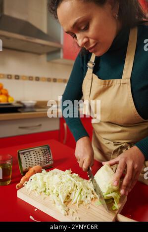 Eine Frau, die eine Schürze trägt, hackt frischen Kohl auf einem Holzbrett in einer warmen Küche. Die Szene weckt ein Gefühl von Gemütlichkeit und gesundem Kochen Stockfoto