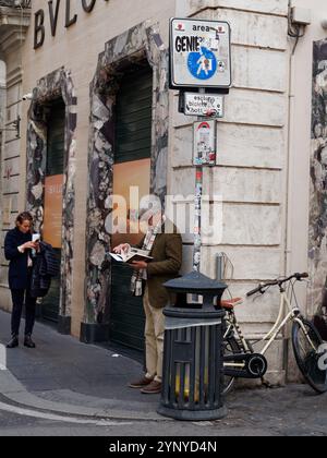 Gut gekleideter reifer Mann mit Brille liest ein Buch an einer Straßenecke neben einem Mülltonne und Fahrrad in Rom, Italien. November 2024 Stockfoto