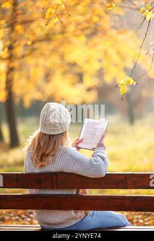 Frau in Strickpullover und Hut sitzt auf der Bank und liest Buch im Herbstpark. Entspannen Sie sich im Freien Stockfoto
