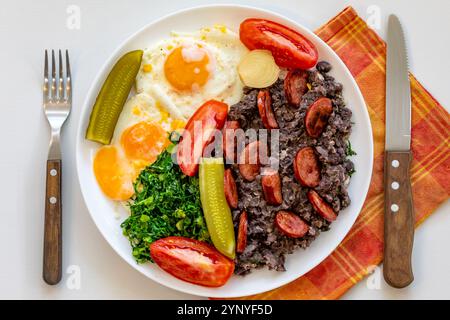 Rustikales, nahrhaftes brasilianisches Essen, Eintopf aus schwarzen Bohnen mit Eiern, gegrillte Würstchen, Gurken und natürliche Gärung, Grünkohl und Gemüse. Echtes Essen Stockfoto