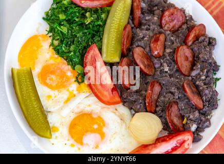 Rustikales, nahrhaftes brasilianisches Essen, Eintopf aus schwarzen Bohnen mit Eiern, gegrillte Würstchen, Gurken und natürliche Gärung, Grünkohl und Gemüse. Echtes Essen Stockfoto