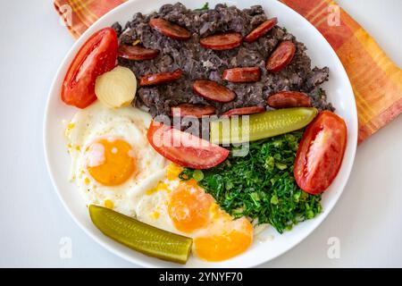 Rustikales, nahrhaftes brasilianisches Essen, Eintopf aus schwarzen Bohnen mit Eiern, gegrillte Würstchen, Gurken und natürliche Gärung, Grünkohl und Gemüse. Echtes Essen Stockfoto