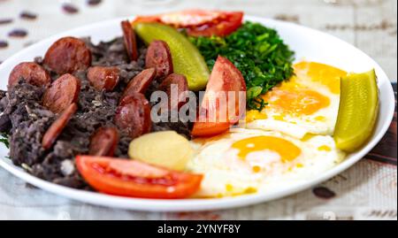 Rustikales, nahrhaftes brasilianisches Essen, Eintopf aus schwarzen Bohnen mit Eiern, gegrillte Würstchen, Gurken und natürliche Gärung, Grünkohl und Gemüse. Echtes Essen Stockfoto