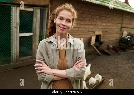 Glückliche junge Farmerin in in grauem Hemd, die mit einem Lächeln gegen Holzhaufen und mehrere Hühner in die Kamera blickt Stockfoto