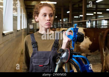 Junge Landarbeiterin mit Melkmaschine in der Hand, die auf einer Viehzucht mit Milchkühen in Reihe geht Stockfoto