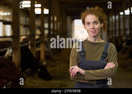Junger Landwirt mit roten, langen lockigen Haaren, der Sie auf einer Viehzucht mit Milchkühen ansieht, während er die Arme an der Brust kreuzt Stockfoto