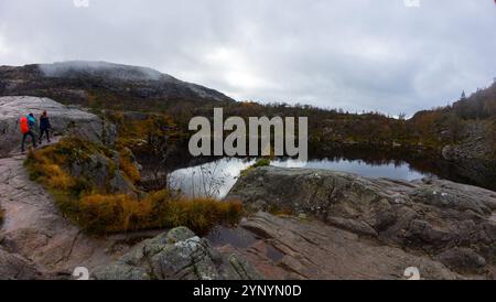 Ein Panoramablick auf einen ruhigen See, umgeben von felsigem Gelände und Herbstlaub. Zwei Wanderer gehen entlang der felsigen Kante, mit nebligen mountai Stockfoto