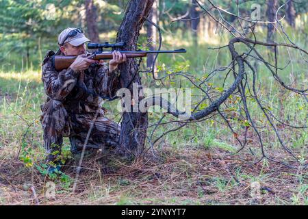 Hunter in Tarnkleidung ist in kniender Position hinter dem Baum und richtet sein Gewehr auf das Spiel, während er durch das Oszilloskop schaut. Seitenansicht. Jagen. Stockfoto