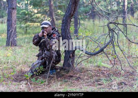 Hunter in Tarnkleidung ist in kniender Position hinter dem Baum und richtet sein Gewehr auf das Spiel, während er durch das Oszilloskop schaut. Vorderansicht. Jagen. Stockfoto