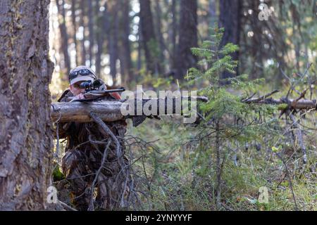 Hunter in Tarnkleidung, zielt auf ein Gewehr und legt es auf einen umgestürzten Baum. Ich schaue durch das Gewehrrohr. Bereit für die Aufnahme. Jagen. Stockfoto