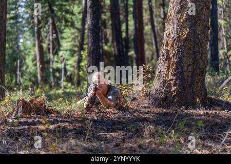 Jäger in Tarnkleidung, der auf dem Boden unter dem Baum liegt und ein Gewehr anvisiert, während er durch das Oszilloskop schaut. Bereit für die Aufnahme. Vorderansicht. Jagen. Stockfoto