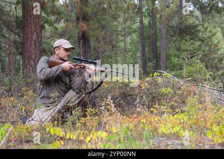 Jagd im Wald. Hunter in kniender Position hält ein Gewehr mit einem Oszilloskop, das auf das Spiel blickt und bereit zum Schießen ist. Seitenansicht. Stockfoto