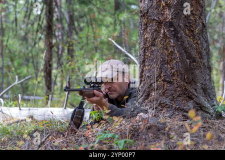 Ein Jäger liegt auf dem Boden hinter einem Baum und richtet sein Gewehr durch das Oszilloskop auf das Spiel, bereit, in einer Waldumgebung zu schießen Stockfoto