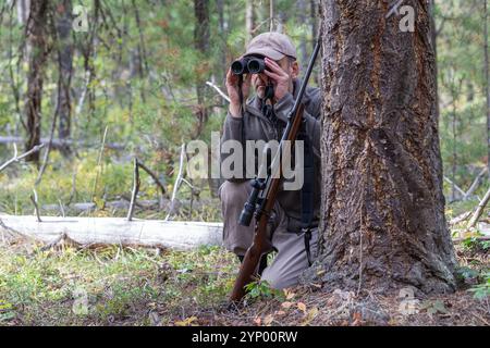 Hunter versteckt sich hinter einem Baum und beobachtet die Umgebung mit einem Fernglas. Ein Gewehr mit dem Zielfernrohr lehnt sich an den Baum vor ihm. Stockfoto