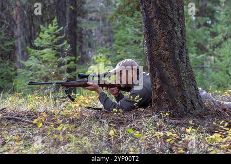 Ein Jäger liegt auf dem Boden hinter einem Baum und richtet sein Gewehr durch das Oszilloskop auf das Spiel, bereit, in einer Waldumgebung zu schießen. Stockfoto