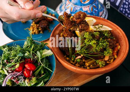 Traditionelle marokkanische Lammtagine mit frischem Salat. Eine Hand schöpft das Gericht und hebt mediterrane Aromen und kulturelle kulinarische Praktiken hervor Stockfoto