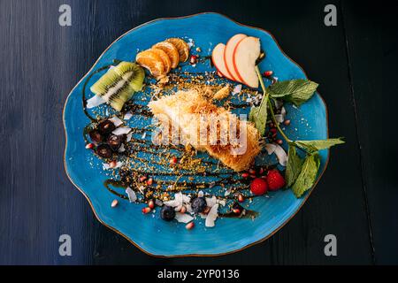 Elegantes Dessert mit Kunafa und frischen Obstscheiben auf blauem Teller. Eine kreative Präsentation von Süßigkeiten aus dem Nahen Osten mit lebhaften Beilagen. Stockfoto