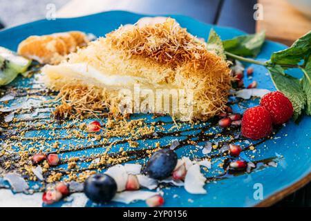 Elegantes Dessert mit Kunafa und frischen Obstscheiben auf blauem Teller. Eine kreative Präsentation von Süßigkeiten aus dem Nahen Osten mit lebhaften Beilagen. Stockfoto