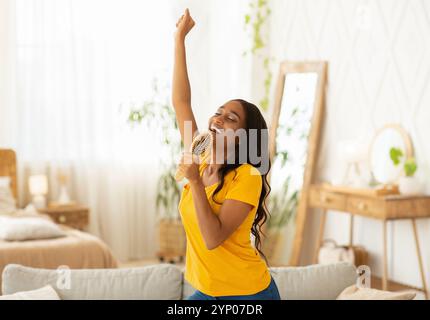 Fröhliche schwarze Frau mit Haarbürste als mic singen ihren Favoriten Gesang und Tanz zu Hause Stockfoto