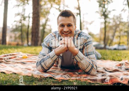 Porträt eines lächelnden, gutaussehenden Mannes mittleren Alters, der auf einer Decke auf grünem Gras im Wald liegt und in die Kamera schaut. Picknick, entspannendes Konzept Stockfoto