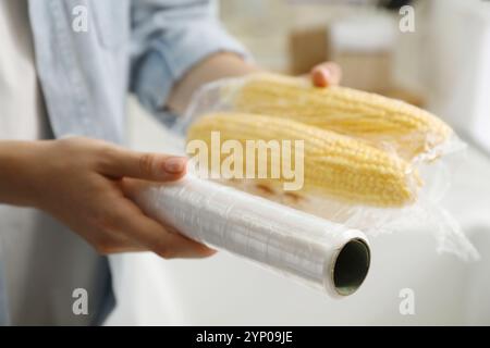 Frau, die Plastikfutterverpackung über Maiskolben in der Küche legt, Nahaufnahme Stockfoto