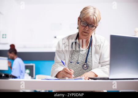 Der ältere Arzt konzentrierte sich auf das Ausfüllen von Rezepten mit offenem Laptop für schnellen Zugriff auf digitale Aufzeichnungen. Pensionierte Ärztin sitzt im Klinikbüro und liest medizinische Notizen auf dem Clipboard. Stockfoto